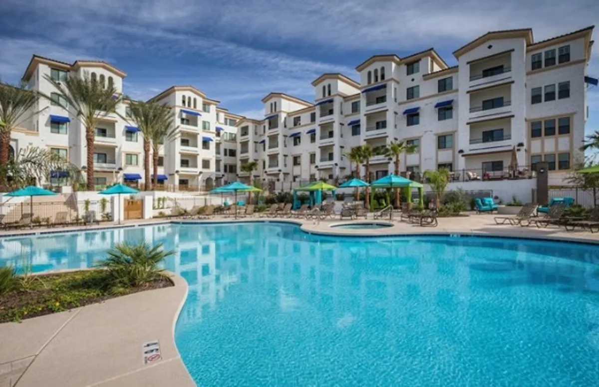 An exterior view that highlights the building's facade, set against a backdrop of palm trees and a sparkling swimming pool with lounge chairs and umbrellas. The scene embodies a sense of arrival and the resort-style atmosphere, hinting at the secure, upscale environment guests experience upon entering.