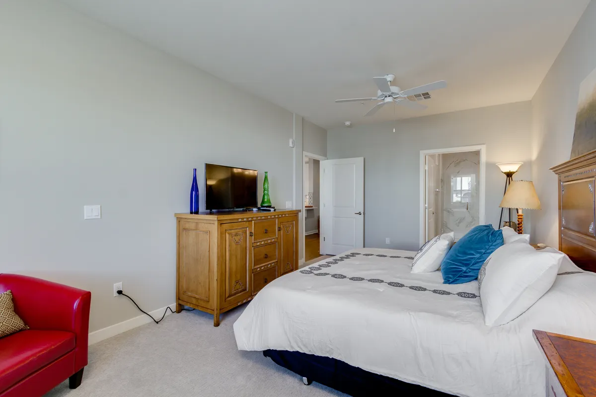 A comfortable bedroom with a king bed dressed in decorative pillows, a red armchair for reading or lounging, a wooden dresser topped with a flat-screen TV, and warm lighting that enhances the cozy ambiance. The layout emphasizes both relaxation and personal entertainment.