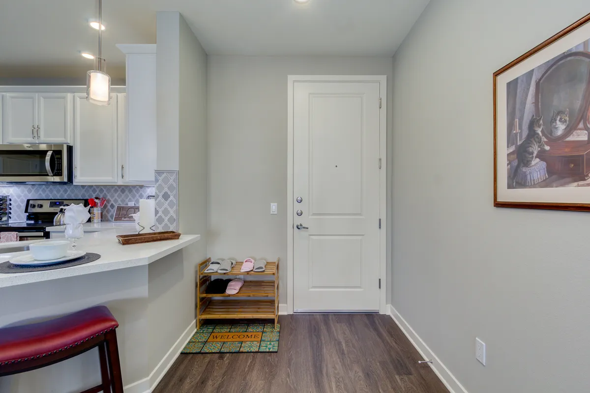 This image captures a stylish interior entryway featuring a modern entry door with a welcome mat, a kitchen bar area visible just beyond, and thoughtfully placed framed artwork. The scene is bathed in inviting, natural light, evoking a sense of comfort and a warm welcome. The layout gives a first impression of entering the home, emphasizing security and arrival.