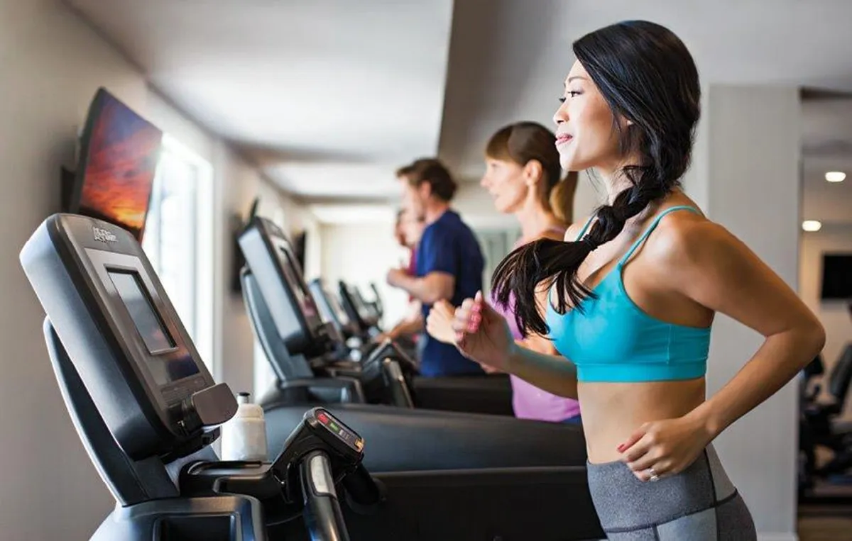 A modern gym with treadmills, people engaged in cardio workouts, and a mounted television screen. The atmosphere is active and motivational, emphasizing health and fitness.