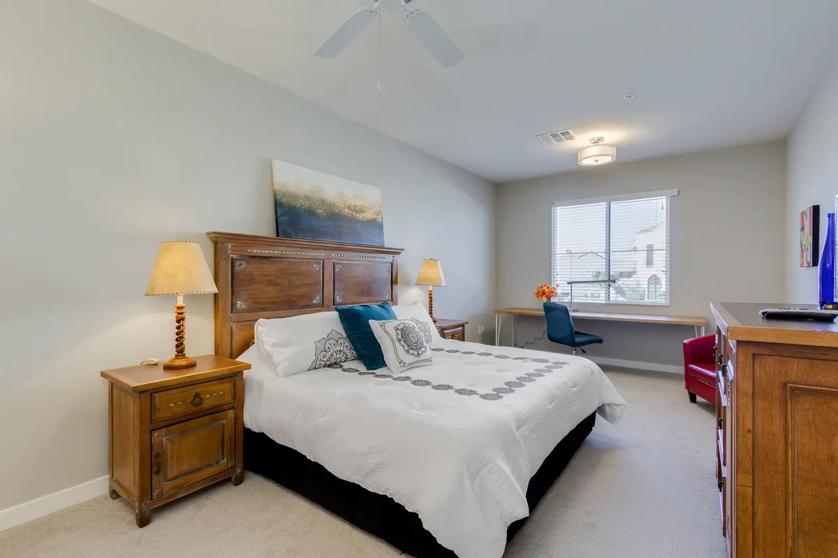 A serene king bedroom featuring a plush bed adorned with decorative pillows, wooden nightstands, a painting above the headboard, and a desk with a chair positioned by a sunlit window. The atmosphere is cozy and tranquil, inviting restful sleep and moments of relaxation or quiet work.