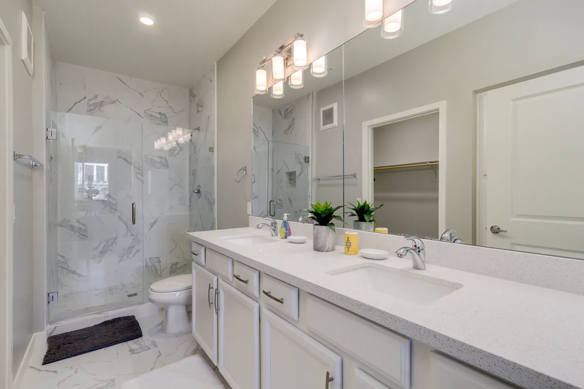 A modern, serene bathroom featuring a double vanity with a large mirror, elegant marble wall tiles, glass shower enclosure, and contemporary light fixtures. The space is bright and luxurious, ideal for self-care routines and relaxation.