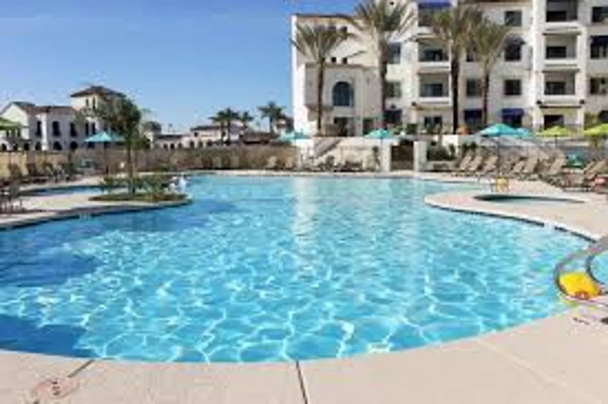 A sunlit pool area surrounded by lounge chairs and palm trees, adjacent to the community's modern buildings. The space is inviting, ideal for unwinding or socializing by the heated pool after a round of golf.