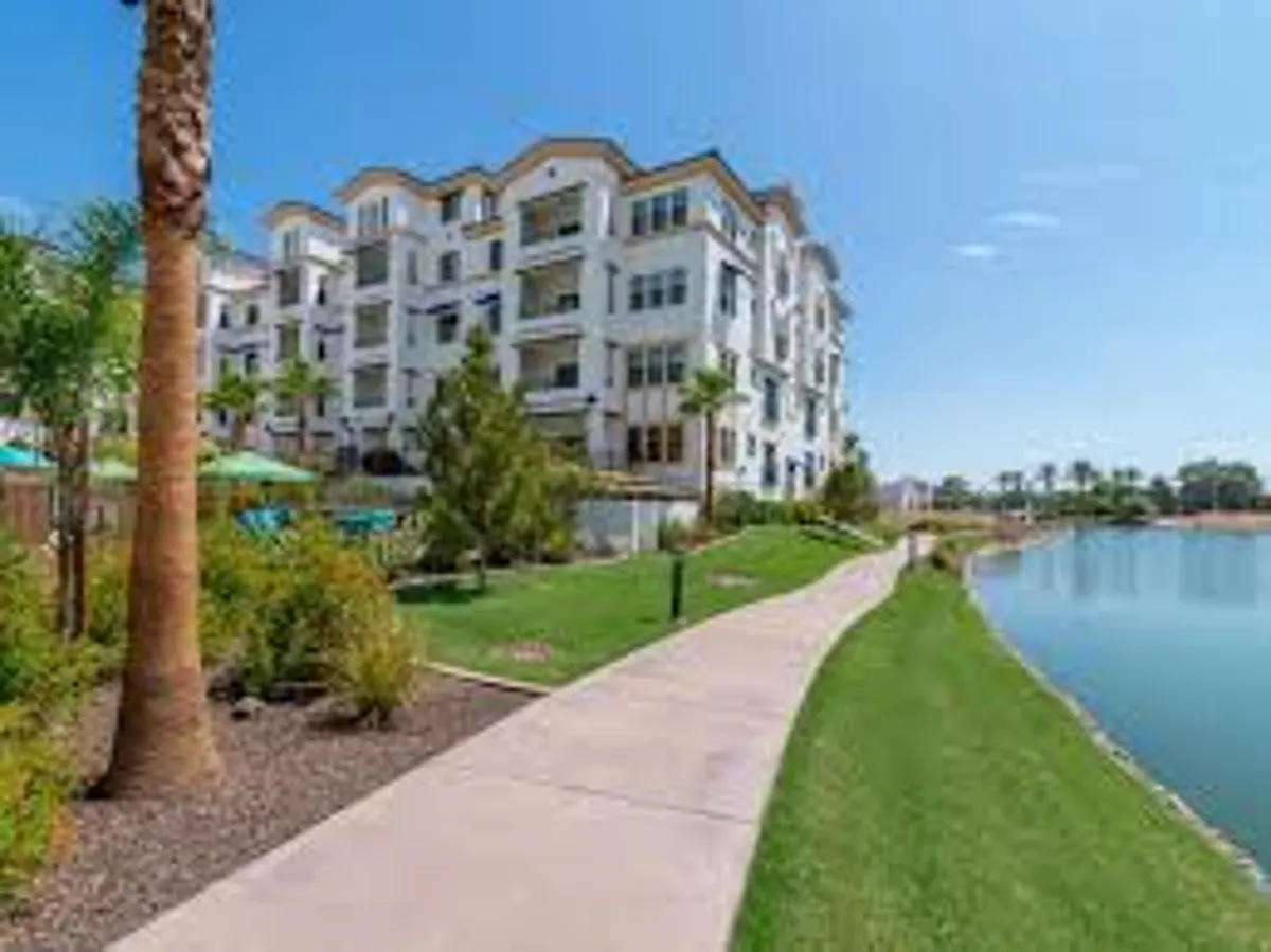 A tranquil exterior shot featuring the modern apartment building of The Cays at Ocotillo. The scene includes a pathway leading toward a serene lake, bordered by palm trees and a lush green lawn, inviting outdoor relaxation and community engagement.