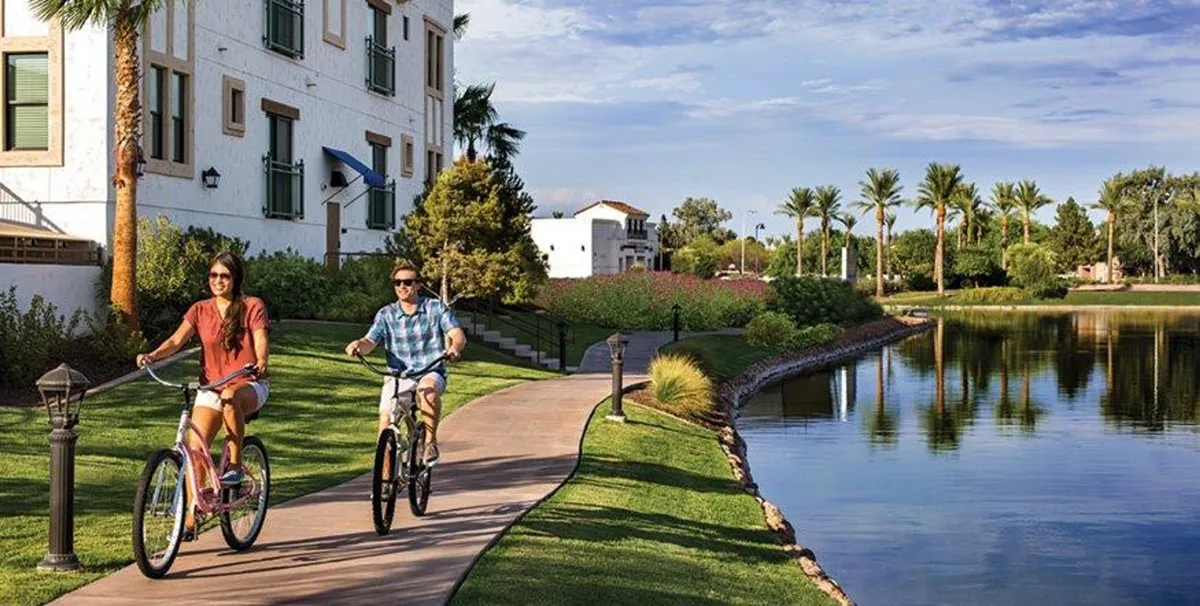 An active, joyful scene showing bicyclists on a path bordering a tranquil lake, with palm trees and community buildings in the background. The mood is lively and relaxed, emphasizing outdoor recreation and the vibrant lifestyle at Ocotillo.